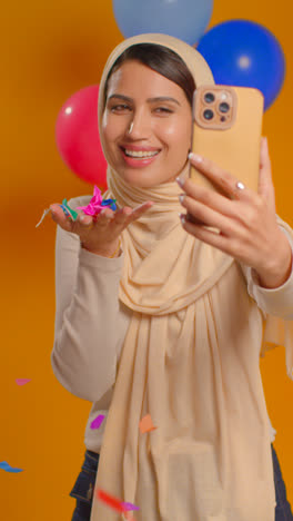 Retrato-De-Estudio-De-Video-Vertical-De-Una-Mujer-Tomando-Selfie-Usando-Hijab-Celebrando-Cumpleaños-Soplando-Confeti-De-Papel-Rodeado-De-Globos